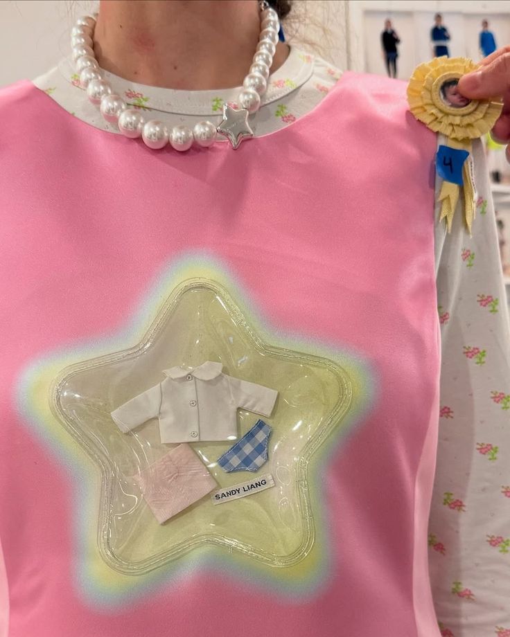 Foto backstage della sfilata di Sandy Liang di autunno-inverno 2025. Vestito rosa con una stella in PVC che presenta all'interno delle miniature di abiti come quelli che indossano le bambole.