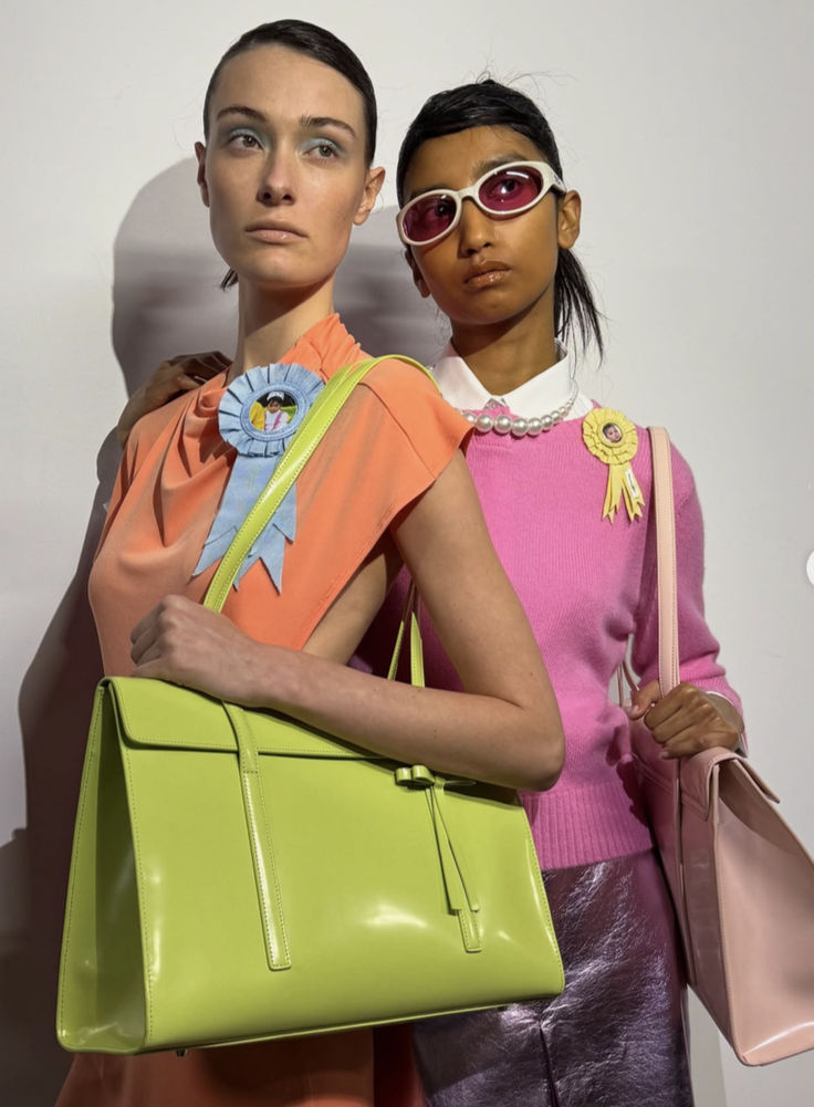 Foto bakcstage dello show di Sandy Liang autunno-inverno 2025. Due modelle indossando colori vivaci come il verde lime, fucsia, arancione, giallo e viola metalizzaro.