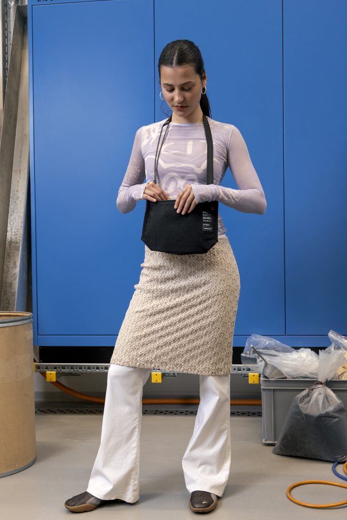 Ragazza con capelli neri indossando una gonna beige sopra pantaloni bianchi, maglietta stampata maniche lunghe e la Mono PA6 Musette di Freitag