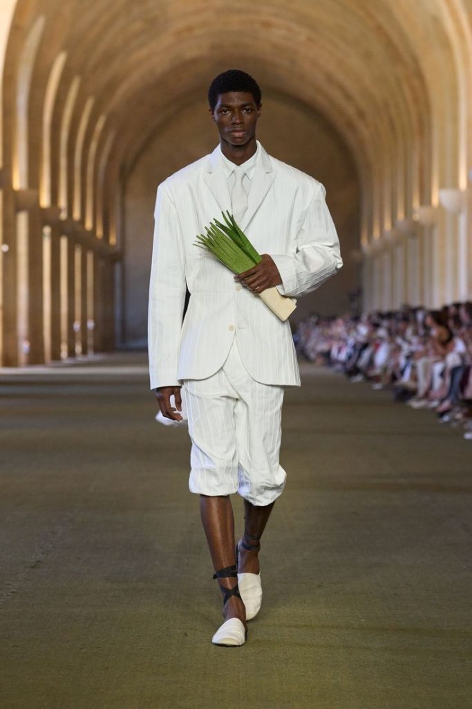 Foto della sfilata di Jacquemus autunno inverno 2026 chiamata Le Paysan in cui vediamo il modello scuro vestito con un completo di giacca e bermuda bianca e ha in mano una borsa a forma di sedano