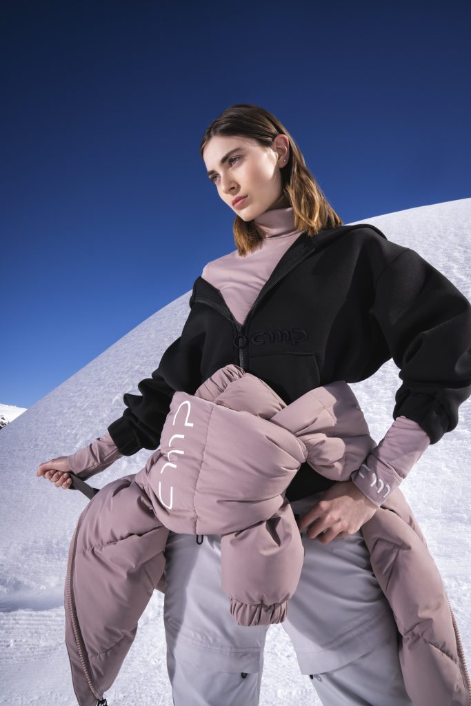 Campagna in un ghiacciao di CMP per presentare la collezione FW25/26. Una ragazza con una felpa con scollo a V nero e un giubbotto rosa antico allacciato alla cintura