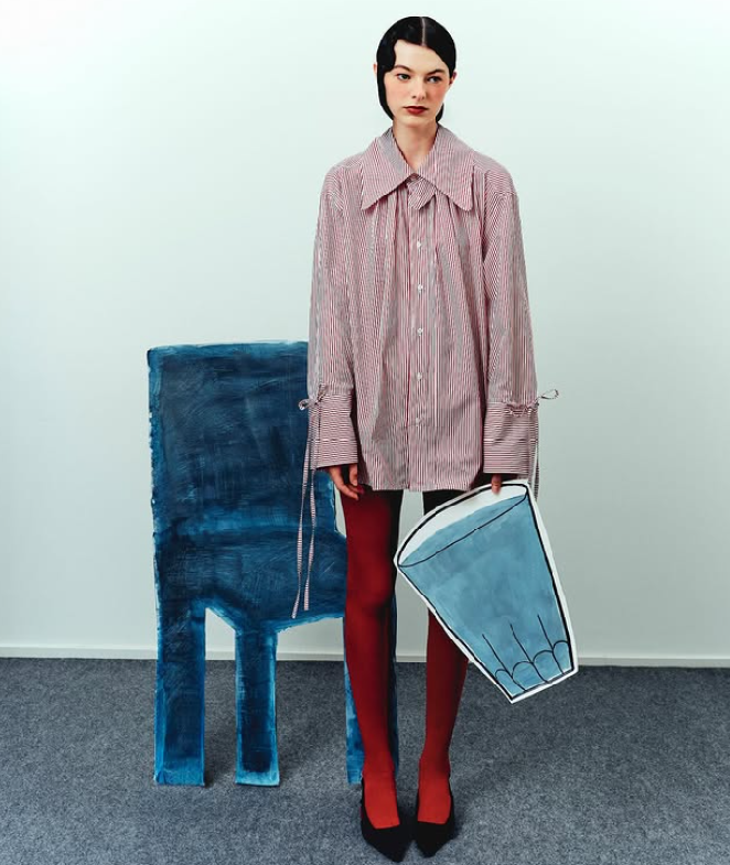 Editoriale di Bombo Studio. Modella in piedi che indossa collant rosse, decollete nere, camicia a righe rosse e bianche e ha degli oggetti in carta in mano