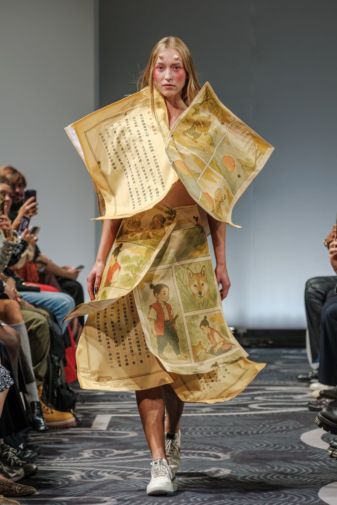 Look 21 della sfilata di APUJAN alla London Fashion Week. La modella indossa un vestito creato a partire da pagine di libri di fiaba giganti tutti ondulati che creano un vestito strutturato e voluminoso