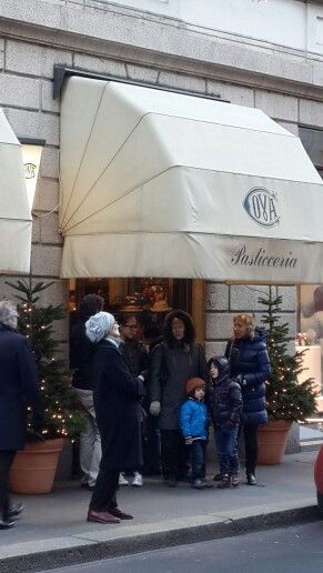 L’immagine mostra l’ingresso della storica Pasticceria Cova, situata in Via Montenapoleone a Milano. La facciata elegante è caratterizzata da una grande tenda beige con il logo “COVA” e la scritta “Pasticceria”. Davanti al negozio, alcune persone, tra cui adulti e bambini, sono raccolte e sembrano in attesa o semplicemente a chiacchierare, con due piccoli alberi di Natale ai lati dell’ingresso che aggiungono un tocco festivo. L’atmosfera riflette il mix di tradizione e vita quotidiana tipico di Montenapoleone.