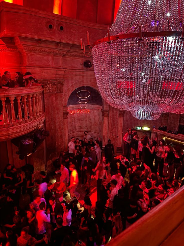 L’immagine mostra l’interno di una elegante discoteca milanese durante una serata affollata. La scena è dominata da un grande e imponente lampadario di cristallo che illumina con riflessi scintillanti la sala. Le pareti in stile classico, con colonne e decorazioni, creano un contrasto con l’atmosfera vivace e moderna della folla. La luce rossa avvolge la scena, mentre un gruppo numeroso di giovani balla e socializza sia al piano inferiore che sulla balconata, rendendo l’ambiente animato e coinvolgente. Sullo sfondo si intravede l’insegna “Cattivardo”.
