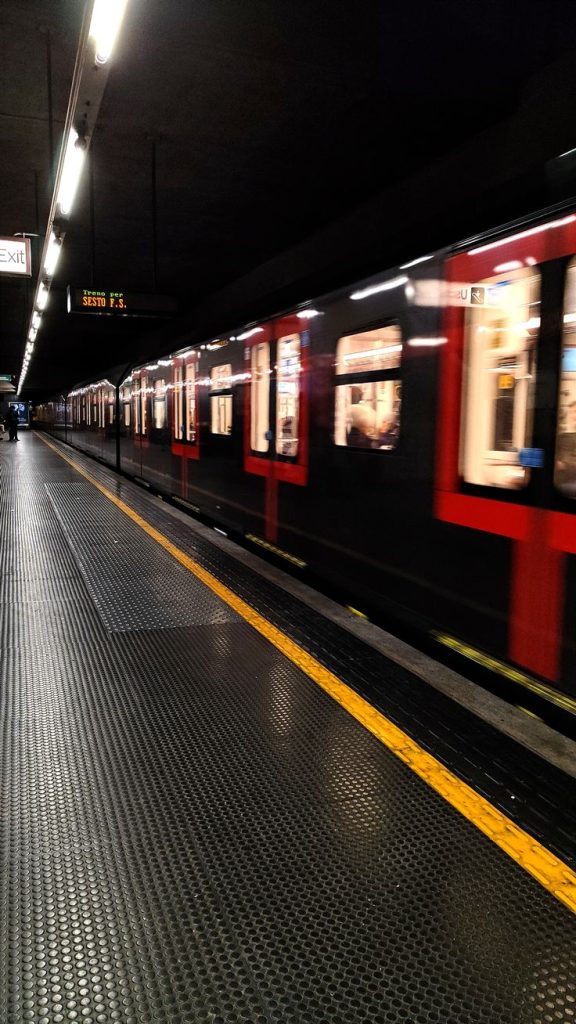 L’immagine mostra una stazione della metropolitana di Milano con un treno in partenza o in arrivo. La banchina è illuminata da luci fluorescenti e ha una pavimentazione con superficie antiscivolo nera, evidenziata da una linea gialla di sicurezza lungo il bordo. Il treno è caratterizzato da porte e finestre bordate di rosso e si muove con velocità, come suggerisce la leggera sfocatura. Sul pannello informativo sopra la banchina si legge la destinazione “Sesto F.S.”. L’atmosfera è tipica di un momento di transito quotidiano nel cuore della città.