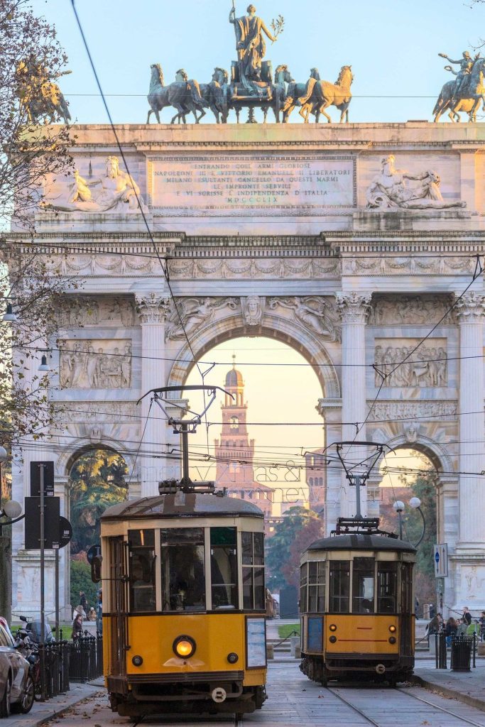 L'immagine mostra un'iconica vista di Milano, con l'Arco della Pace sullo sfondo. Due tram gialli si incrociano sotto l'arco, mentre si intravede una torre (probabilmente la Torre Branca) oltre l'arco stesso. La scena è ambientata in un pomeriggio luminoso, con una luce calda che illumina i dettagli architettonici dell'arco e degli edifici circostanti. L'atmosfera è vivace, con persone che camminano lungo la strada, mentre le linee dei tram e dei fili elettrici aggiungono un tocco di modernità alla storicità del luogo.