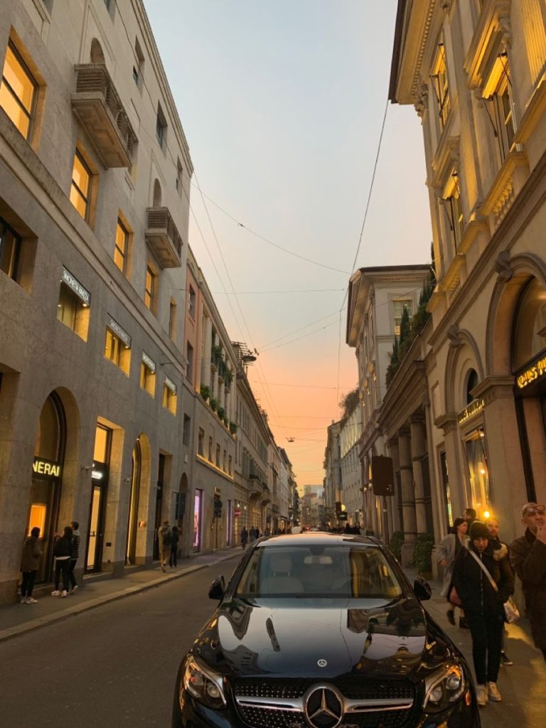 L'immagine mostra Via Montenapoleone in un'atmosfera serale, con il cielo che sfuma nei toni del rosa e dell'arancione al tramonto. In primo piano si vede un'auto Mercedes nera parcheggiata al centro della strada, mentre sul lato destro alcune persone camminano. Gli edifici sono eleganti, con vetrine di negozi di lusso e dettagli architettonici raffinati. La scena trasmette una sensazione di tranquillità e classe.