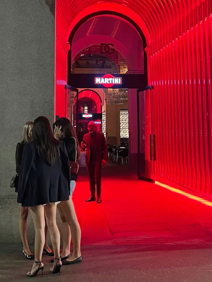 L'immagine mostra un'atmosfera notturna vivace e moderna. In primo piano ci sono alcune persone, probabilmente giovani, che stanno chiacchierando sotto una luce rossa intensa, che illumina l'intera scena. La zona è caratterizzata da un'illuminazione d'atmosfera, con le luci rosse provenienti da una struttura architettonica che crea un tunnel luminoso. Sullo sfondo, si vede un'insegna del famoso marchio MARTINI, suggerendo che si tratta di una zona di intrattenimento o di un locale alla moda. L'ambiente sembra essere elegante e trendy, con una combinazione di persone vestite in modo formale e informale.