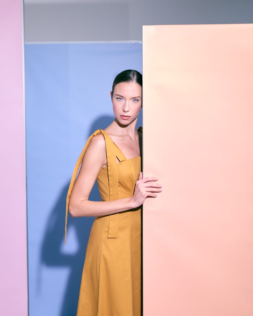 Donna in abito midi color senape, linee pulite e dettagli sartoriali sulle spalle.
Atmosfera grafica e sofisticata, con contrasti soft tra pastelli e posa elegante.