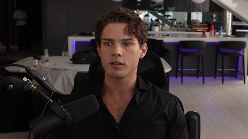 Clavicular durante una livestream in un appartamento moderno, seduto davanti a un microfono mentre parla di looksmaxing e cultura internet.