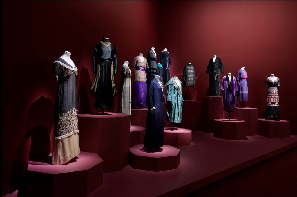 Esposizione di abiti storici su manichini, allestiti su pedane davanti a pareti rosso bordeaux in una mostra di moda.
