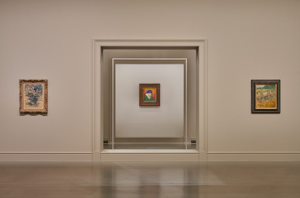 Sala museale con tre dipinti di Vincent van Gogh esposti su pareti beige, con un autoritratto al centro.
