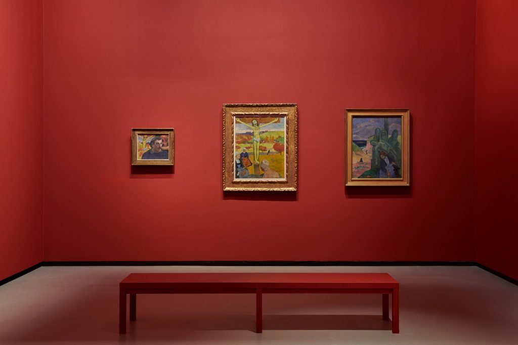 Tre dipinti di Paul Gauguin esposti su una parete rosso intenso, con una panca al centro della sala museale.